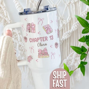 May include: White tumbler with a lid and handle, decorated with pink gift boxes, bows, and coffee cups. The text "CHAPTER 13 Olivia" is displayed on the tumbler. A straw is inserted into the lid.