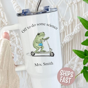May include: White tumbler with a lid and handle, featuring a cartoon frog riding a scooter and holding a beaker. Text reads "Off to do some science" and "Mrs. Smith". A pink sticker says "Ships Fast".