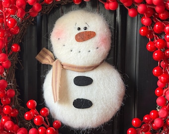 May include: A handmade snowman decoration with a felt body, a carrot nose, and a striped scarf. The snowman is surrounded by a wreath of red berries. The wreath is a festive holiday decoration.