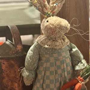 May include: A decorative Easter bunny doll with a cream-colored, textured body and floral patterned ears. The bunny wears a light green and white checkered dress with a lace collar and holds two orange carrot decorations. A rustic Easter decoration.