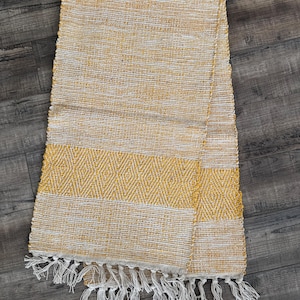 May include: A woven rectangular throw blanket in shades of yellow and white, with a diamond pattern in the center and white fringe along the bottom edge. The blanket is made of a textured material.