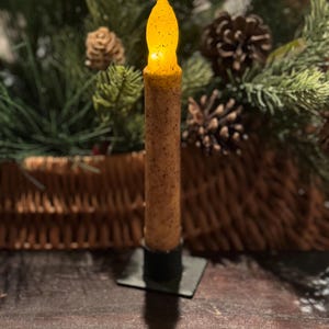 May include: A battery-operated, flameless candle with a flickering amber LED flame. The candle has a textured, brown wax-like appearance and sits on a black square base. The background features pine branches, pine cones, and a woven basket.