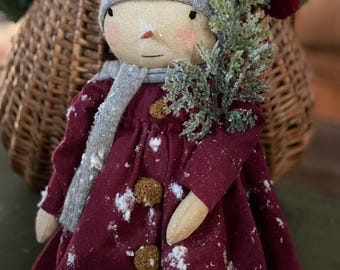 May include: A handcrafted snowman doll wearing a gray knit cap with a red pom-pom, a gray scarf, and a burgundy coat with three tan pom-pom buttons. The doll holds a small evergreen branch, and is dusted with white accents, suggesting a snowy scene.