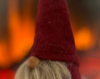 May include: A small, handmade gnome figurine with a burgundy felt hat and a long, fluffy, light-colored beard. The gnome has a small, round nose. The background is blurred, with warm orange and red tones, suggesting a fireplace or warm setting.