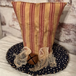 May include: A handmade patriotic hat with a red and tan striped fabric top and a blue base with white stars. A brown bell is attached to the hat.