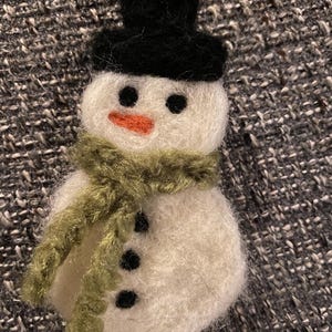 May include: A white felt snowman ornament with a black top hat and a green scarf. The snowman has black eyes, an orange nose, and three black buttons.
