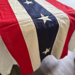 May include: A patriotic table runner with a design featuring red and white stripes, and a blue section with white stars. The runner is made of fabric and is likely used for decorative purposes during holidays or special events.