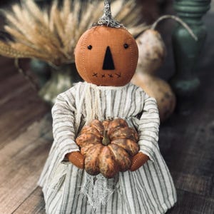 May include: A fabric doll with a round orange pumpkin head featuring a black triangular nose, two small black eyes, and a stitched mouth. The doll wears a long-sleeved dress with vertical grey and white stripes and holds a small, textured pumpkin.