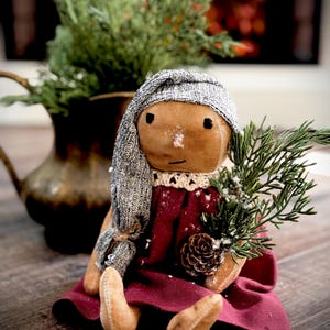 May include: A handmade doll with a tan face, black eyes, and a gray knitted hood. The doll wears a burgundy dress and holds a sprig of greenery and a pinecone. A decorative pitcher with greenery is in the background.