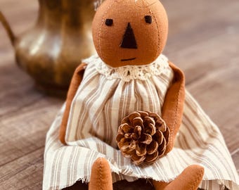 Puede incluir: Muñeca de calabaza hecha a mano con cuerpo marrón, ojos negros y nariz triangular. La muñeca viste un vestido crema con rayas verticales y cuello de encaje. Una piña descansa en su regazo, y hay dos más cerca.