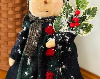 May include: A fabric snowman doll wearing a dark green plaid dress and a gray knit hat and scarf. The doll holds a sprig of holly with red berries. The doll has a tan face with black eyes and a stitched smile. Snow-like accents are scattered on the doll.