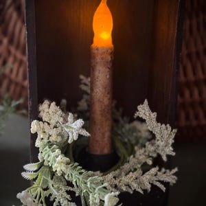 May include: A battery-operated candle with a flickering amber flame sits inside a dark wooden box. The candle is surrounded by a green wreath with white and green floral accents. The candle has a textured, brown appearance.