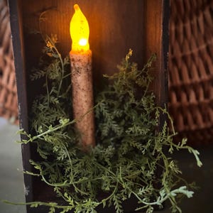 May include: A battery-operated candle with a flickering yellow flame sits inside a dark wooden box. The candle is surrounded by green leafy vines, creating a rustic and cozy aesthetic. The candle has a textured, aged appearance.