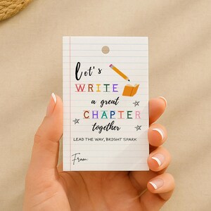 May include: A white card with a lined paper design, featuring the text "Let's WRITE a great CHAPTER together." The card includes a pencil and open book graphic, with the words in various colors. The text "LEAD THE WAY, BRIGHT SPARK" is also present.
