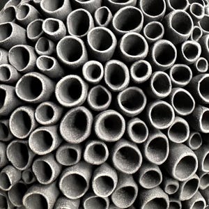 May include: Close-up of a textured, gray ceramic sculpture. The artwork features numerous cylindrical openings of varying sizes, creating a honeycomb or coral-like pattern. The openings are dark, contrasting with the lighter gray exterior.