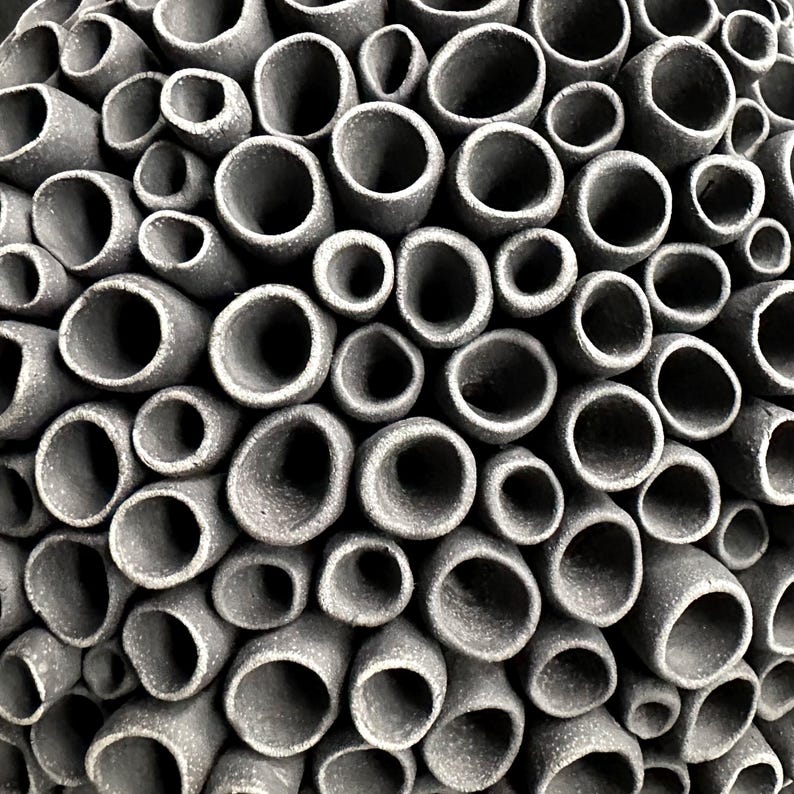 May include: Close-up of a textured, gray ceramic sculpture. The artwork features numerous cylindrical openings of varying sizes, creating a honeycomb or coral-like pattern. The openings are dark, contrasting with the lighter gray exterior.