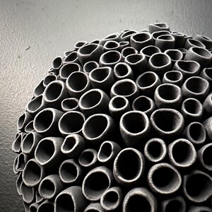 May include: A spherical, dark gray ceramic sculpture with a textured surface. The sculpture is composed of numerous cylindrical openings of varying sizes, creating a honeycomb-like pattern. The artwork is set against a neutral gray background.