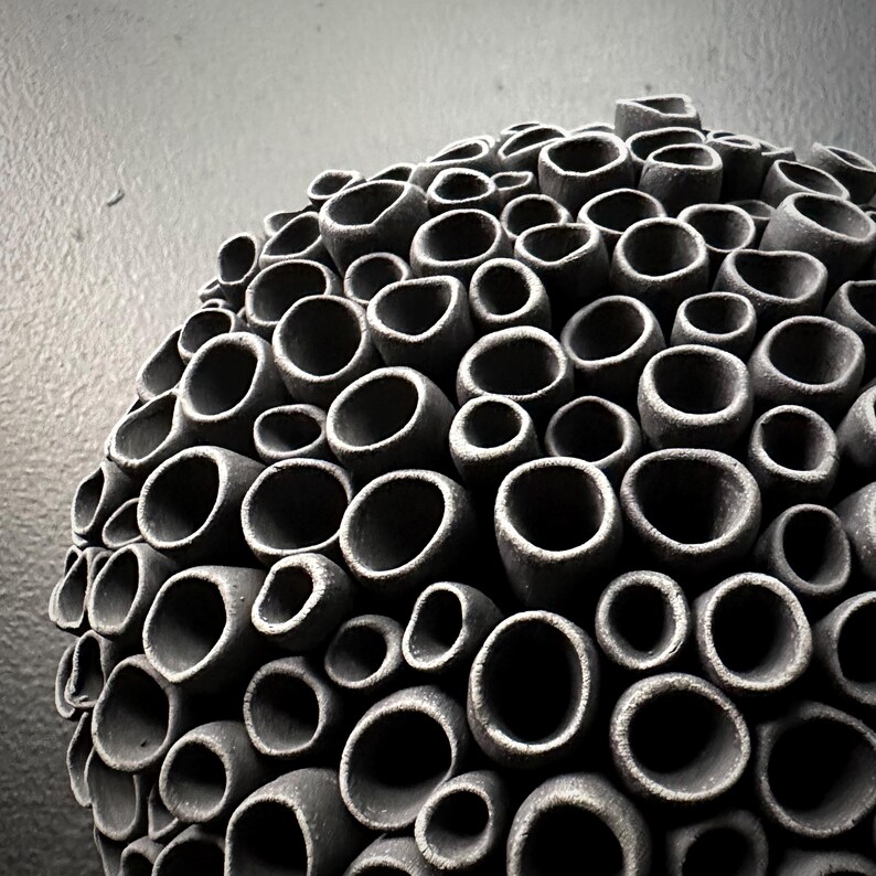 May include: A spherical, dark gray ceramic sculpture with a textured surface. The sculpture is composed of numerous cylindrical openings of varying sizes, creating a honeycomb-like pattern. The artwork is set against a neutral gray background.