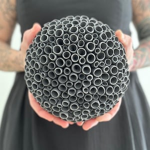 May include: A spherical sculpture composed of numerous gray, cylindrical tubes, creating a textured, porous surface. The artwork is held in both hands, showcasing its unique, organic form. The sculpture's design suggests a modern, abstract aesthetic.