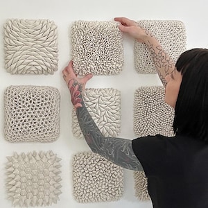 May include: A wall display of nine white ceramic tiles with different textured surfaces. The tiles are arranged in a grid pattern and are all the same size. The tiles are made of a smooth, white ceramic and have a variety of textures, including raised dots, ridges, and swirls.