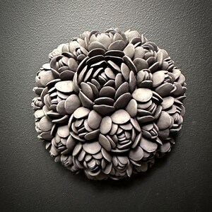 May include: A circular wall sculpture made of gray ceramic, resembling a cluster of stylized flowers. The sculpture is composed of numerous overlapping petals, creating a textured and organic appearance.