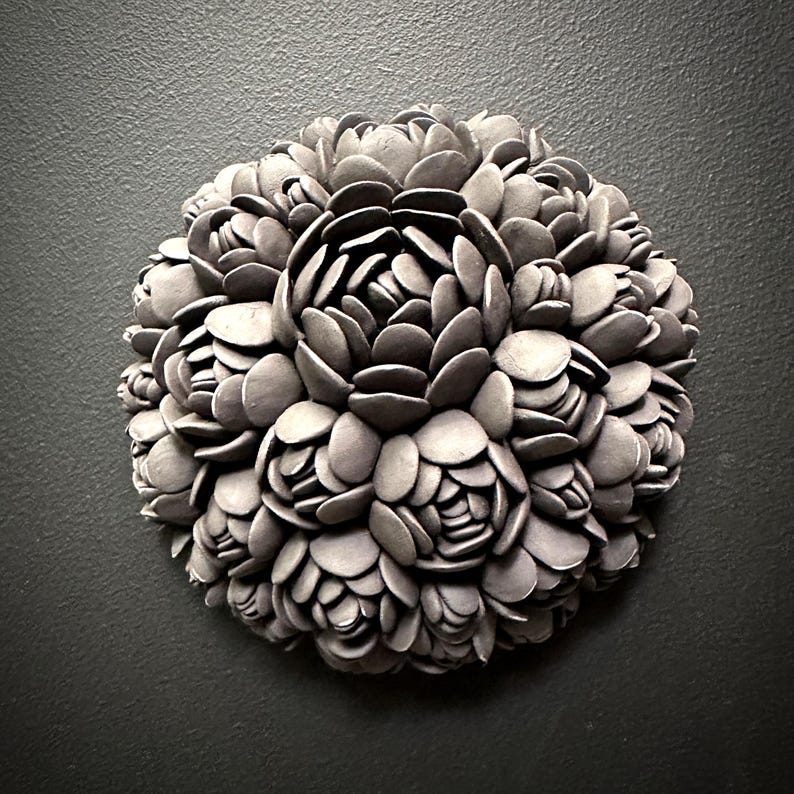 May include: A circular wall sculpture made of gray ceramic, resembling a cluster of stylized flowers. The sculpture is composed of numerous overlapping petals, creating a textured and organic appearance.