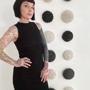 May include: A woman wearing a black sleeveless dress with floral tattoos on her arms stands in front of a white wall with a row of nine round, textured ceramic wall sculptures. The sculptures are in shades of white and gray.
