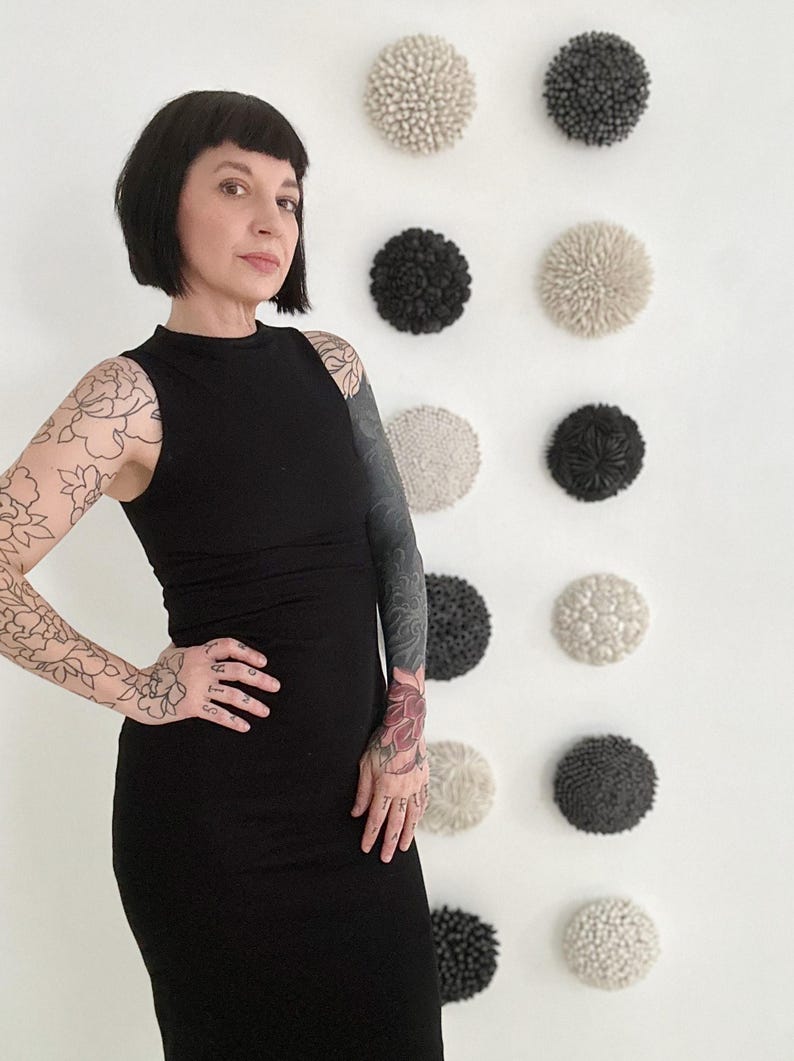 May include: A woman wearing a black sleeveless dress with floral tattoos on her arms stands in front of a white wall with a row of nine round, textured ceramic wall sculptures. The sculptures are in shades of white and gray.