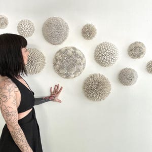 Könnte beinhalten: Eine weiße Wand mit einer Sammlung weißer Keramik-Wandskulpturen in verschiedenen Formen und Größen. Die Skulpturen sind in einem zufälligen Muster angeordnet. Eine Person mit Tätowierungen steht vor der Wand.
