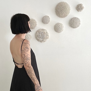 May include: A person wearing a black dress with a floral tattoo on their arm, standing in front of a white wall with several white ceramic wall sculptures.