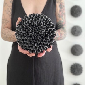 May include: A dark gray ceramic sculpture of a flower with a round shape and a spiral pattern. The sculpture is made of many small, rolled pieces of clay.