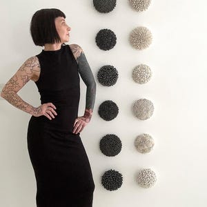 May include: A woman wearing a black dress and black heels stands in front of a white wall with a row of black and white ceramic wall sculptures.