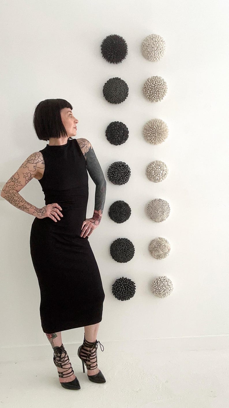 May include: A woman wearing a black dress and black heels stands in front of a white wall with a row of black and white ceramic wall sculptures.