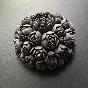 May include: A black ceramic sculpture of a flower with a round shape. The sculpture is made up of many small, overlapping petals.