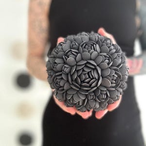 May include: A black and gray floral arrangement made of small, round, petal-like pieces. The arrangement is spherical in shape and appears to be made of a ceramic material.