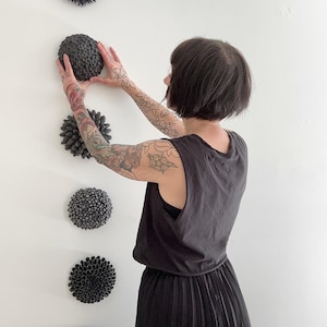 May include: A collection of five dark gray, textured, spherical wall art pieces. The pieces are arranged vertically on a white wall. The art pieces have various textures, including spiky and rounded designs. The overall aesthetic is modern and minimalist.