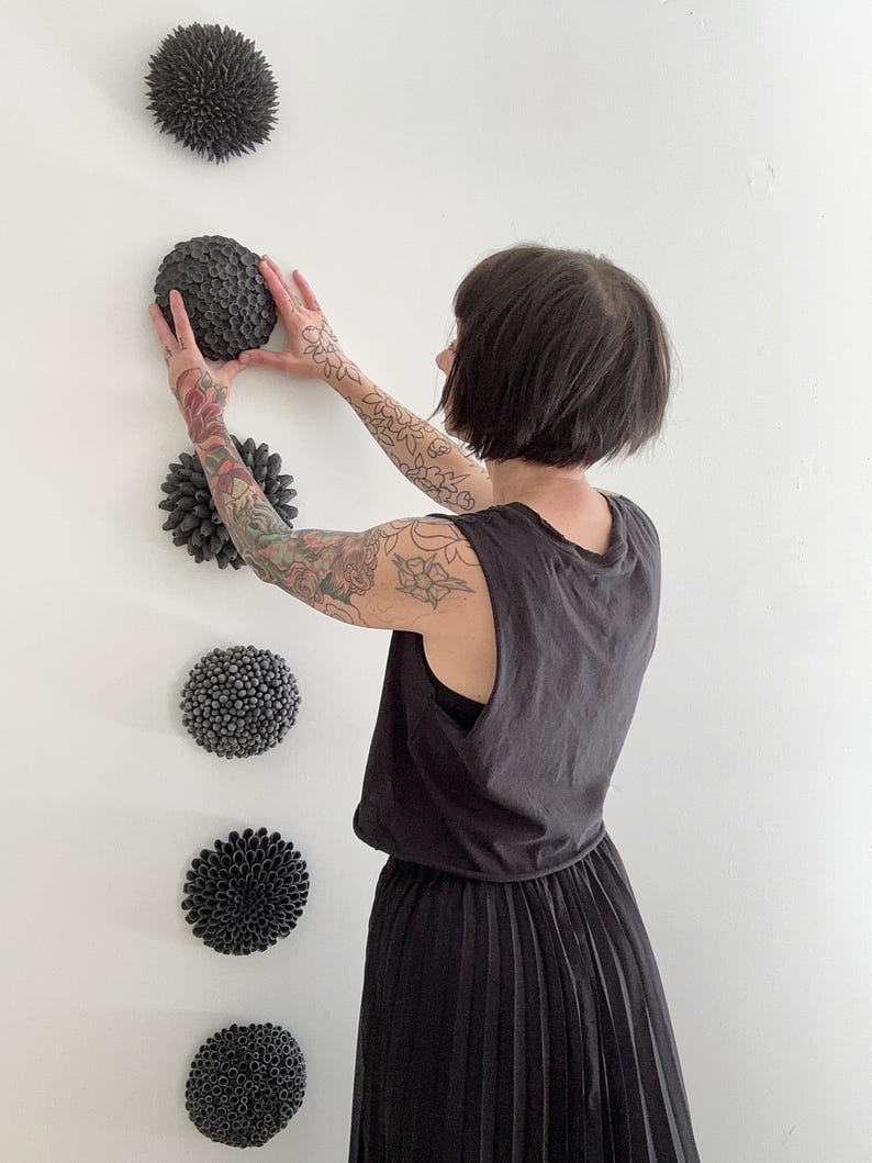 May include: A collection of five dark gray, textured, spherical wall art pieces. The pieces are arranged vertically on a white wall. The art pieces have various textures, including spiky and rounded designs. The overall aesthetic is modern and minimalist.