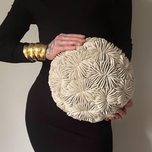 May include: A cream-colored, spherical decorative object with a textured, floral-like pattern. The object is held in front of a person wearing a black dress and gold cuff bracelets.