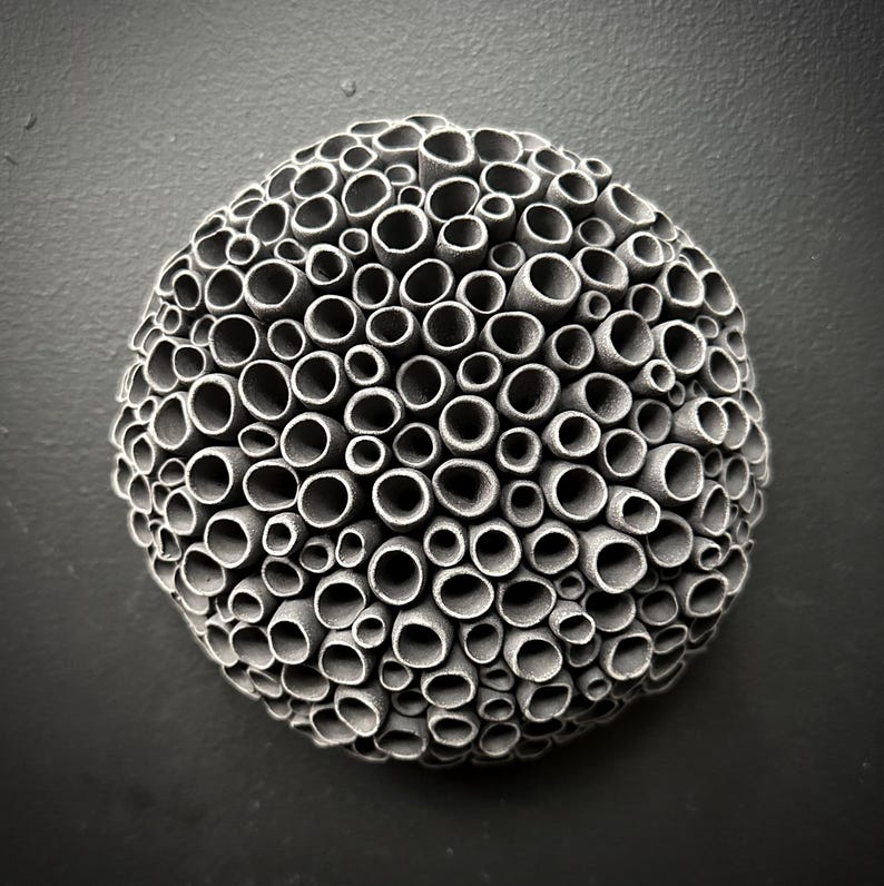 May include: A gray, spherical wall sculpture composed of numerous small, cylindrical tubes. The tubes are arranged in a dense, honeycomb-like pattern, creating a textured surface. The sculpture is mounted on a dark gray wall.