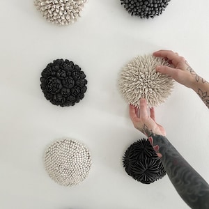 May include: A collection of ceramic wall art pieces in various shapes and textures. The pieces are in shades of black, white, and gray, with some resembling flowers or sea urchins. One piece is being held by a person with tattoos.