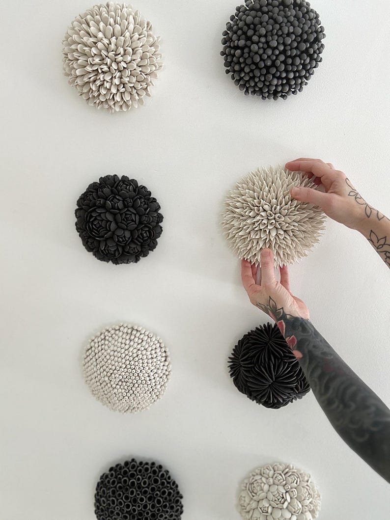 May include: A collection of ceramic wall art pieces in various shapes and textures. The pieces are in shades of black, white, and gray, with some resembling flowers or sea urchins. One piece is being held by a person with tattoos.