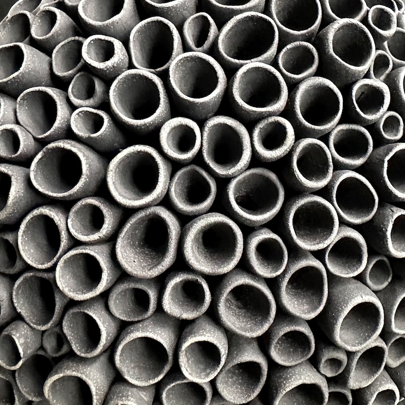 May include: Close-up of a textured, gray ceramic sculpture. The artwork features numerous cylindrical openings of varying sizes, creating a honeycomb or coral-like pattern. The openings are dark, contrasting with the lighter gray exterior.