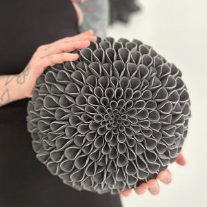 May include: A dark gray, spherical decorative object with a layered petal design, resembling a large flower. The object is held by hands, and the background is a plain white wall. The overall aesthetic is modern and artistic.