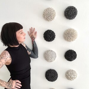 May include: A wall display of nine round ceramic sculptures. The sculptures are arranged in three rows of three. Each sculpture is a different design, with some featuring raised dots, others with raised flowers, and others with raised spikes. The sculptures are all in shades of black and white.