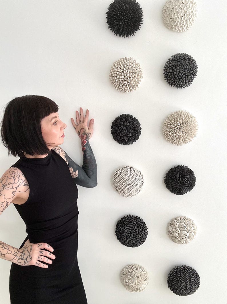 May include: A wall display of nine round ceramic sculptures. The sculptures are arranged in three rows of three. Each sculpture is a different design, with some featuring raised dots, others with raised flowers, and others with raised spikes. The sculptures are all in shades of black and white.