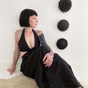 May include: Black halter jumpsuit with a cutout design. The model is sitting on a cream-colored rug. Decorative black wall art in the shape of flowers is mounted on the white wall behind the model.
