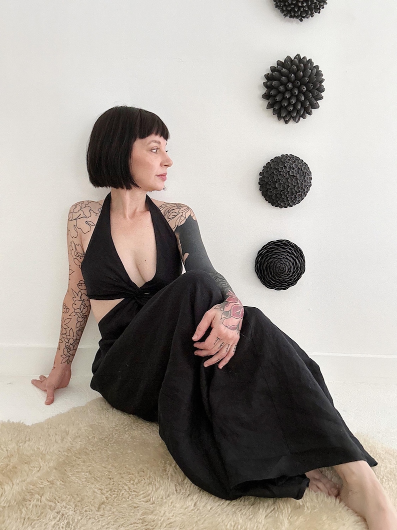 May include: Black halter jumpsuit with a cutout design. The model is sitting on a cream-colored rug. Decorative black wall art in the shape of flowers is mounted on the white wall behind the model.