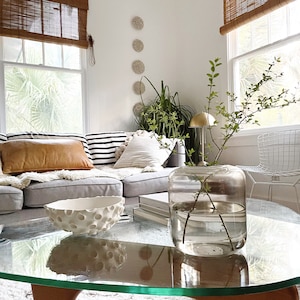 May include: A living room scene featuring a glass coffee table with a decorative white bowl and a large glass vase holding green branches. A gray sectional sofa is adorned with pillows in various textures and patterns, including a tan leather pillow.