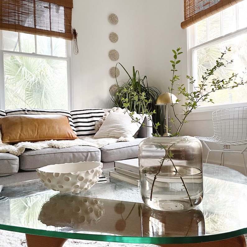 May include: A living room scene featuring a glass coffee table with a decorative white bowl and a large glass vase holding green branches. A gray sectional sofa is adorned with pillows in various textures and patterns, including a tan leather pillow.