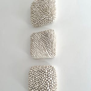 May include: Three white ceramic wall sculptures with different textured surfaces. The top sculpture has a spiky texture, the middle sculpture has a wavy texture, and the bottom sculpture has a round, pebble-like texture.